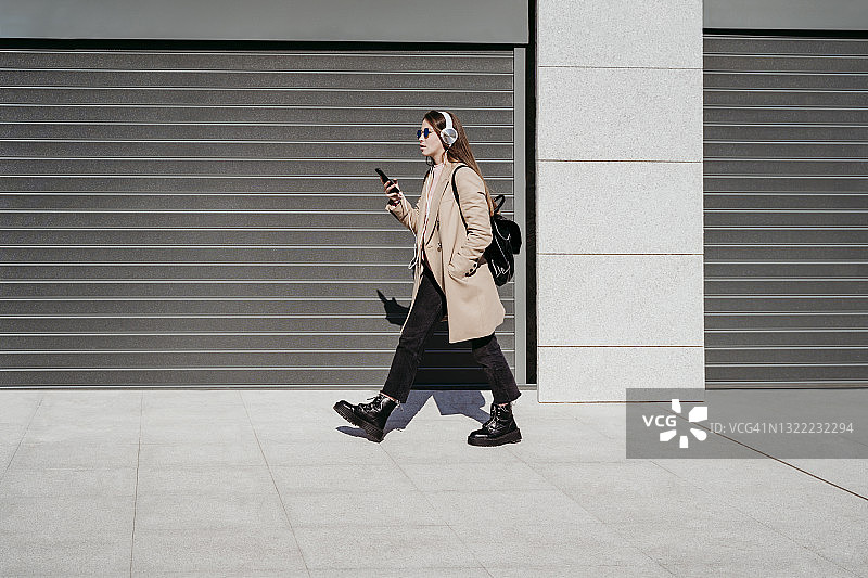 年轻女子戴着耳机和手机走在人行道上图片素材
