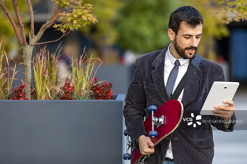 一位穿着西装的年轻英俊男子正在城市街道上玩着滑板冲向办公室，同时用他的数字平板电脑交流。图片素材