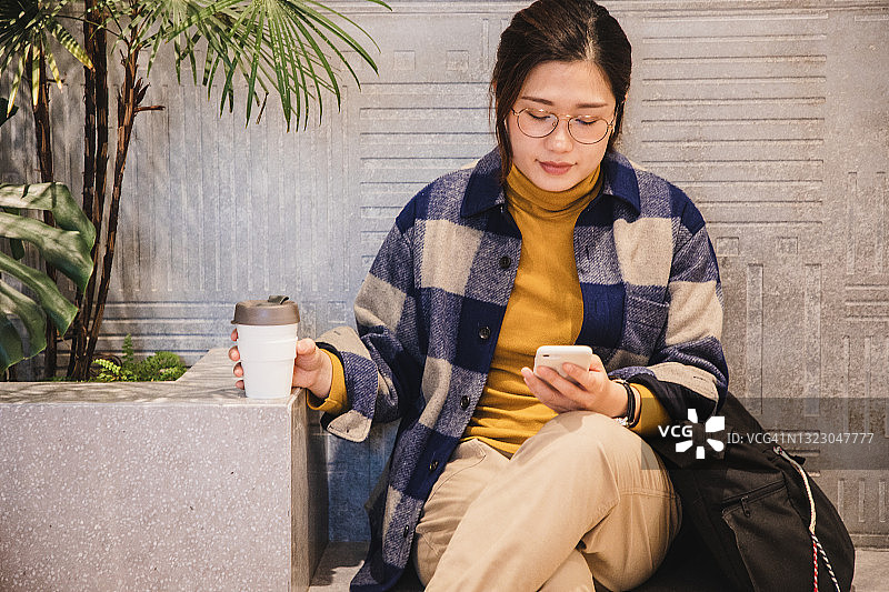 亚洲女性坐着使用电话，用可重复使用的咖啡杯喝咖啡图片素材