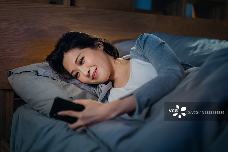 微笑的年轻亚洲女人在睡觉前躺在床上用智能手机和朋友/家人视频通话，在舒适的家里度过一个寒冷的夜晚图片素材