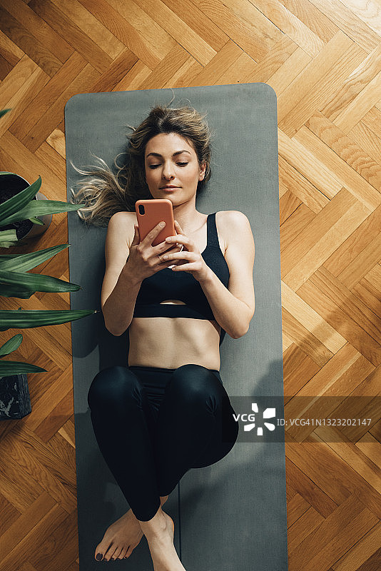 年轻女子躺在瑜伽垫上，从上面使用她的智能手机图片素材