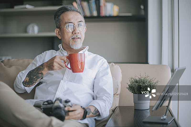 亚洲华人退休老人放松在客厅喝咖啡看别处拿着相机坐在沙发上图片素材
