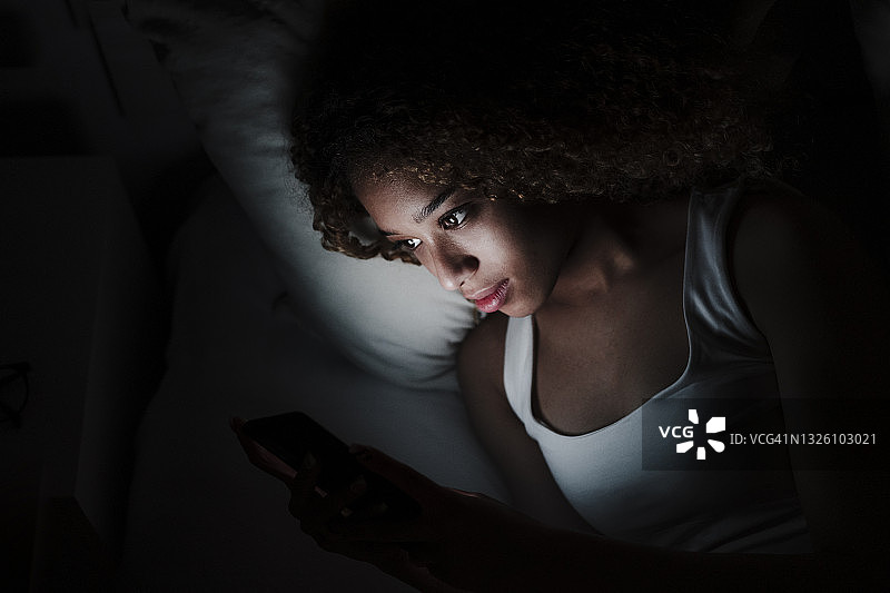 一名女子躺在家中暗室的床上使用智能手机图片素材