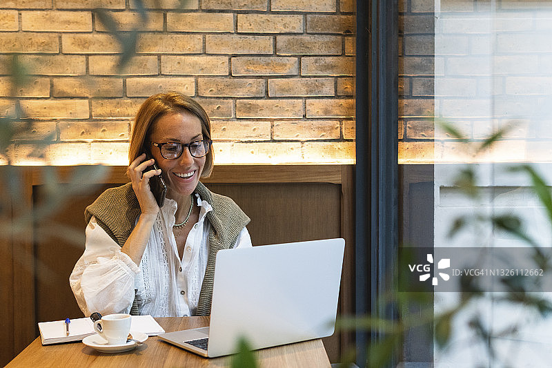 微笑的女人一边用笔记本电脑一边在咖啡店打电话图片素材