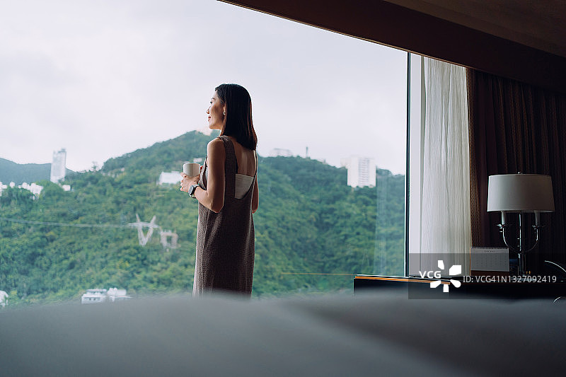 年轻的亚洲女人喝着咖啡，在舒适的家里靠窗放松，看着窗外美丽的风景在早晨。生活方式与片刻的平静和宁静图片素材