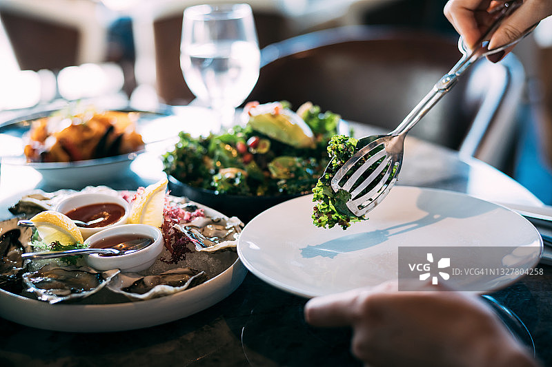 一个女人的手提供新鲜的健康沙拉到一个盘子，而享受美味的一餐在餐厅。外出就餐的生活方式图片素材