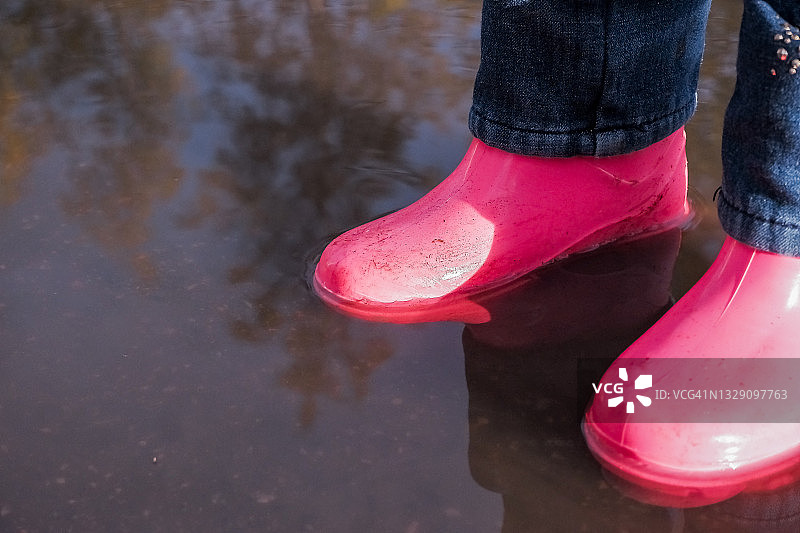 穿着雨靴站在水坑里的孩子的腿。孩子们在秋天。一个穿着粉色橡胶靴子的小女孩的腿在秋天走路的特写。孩子们在户外玩得很开心。小女孩的防水靴。图片素材