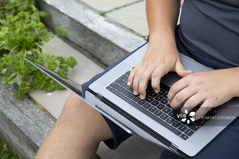 一个十几岁的男孩坐在花园庭院的台阶上，在膝上的笔记本电脑键盘上打字的手的特写图片素材