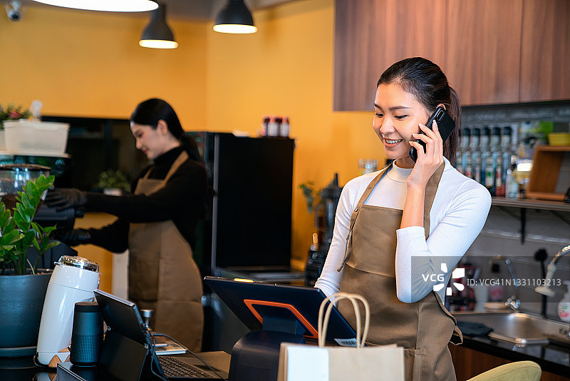 在咖啡店里一边用笔记本电脑工作一边打电话的微笑女人(销售点)。后台的咖啡师正在煮咖啡。图片素材