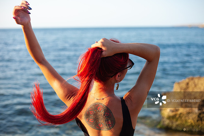 在海边的沙滩上，一位有着长长的红色头发、背上有纹身的年轻女子正在欣赏美丽的日落。图片素材