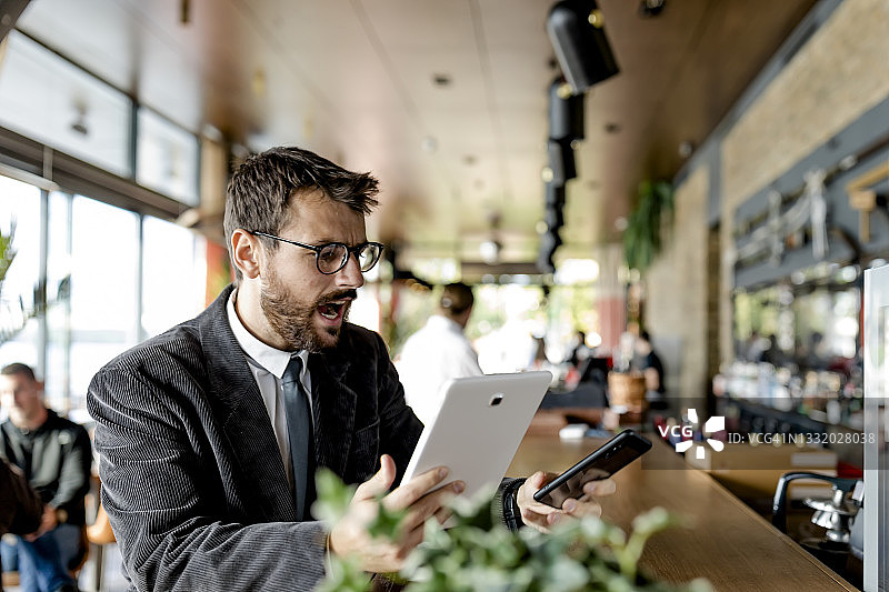 一位年轻快乐的商人坐在现代自助餐厅里，拿着数码平板电脑，用手机发信息。图片素材