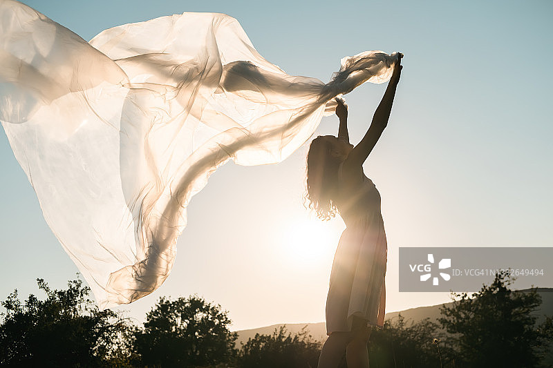 一个年轻女子站在草地上，手里拿着在风中飘动的半透明塑料玻璃纸图片素材