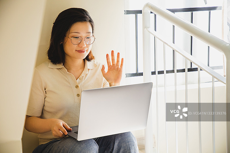 一个亚洲女人坐在楼梯上向手提电脑挥手图片素材