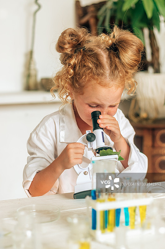 小女孩穿着白色制服，戴着防护眼镜，在一个家庭实验室里看着显微镜。回到学校的概念。年轻的科学家。自然科学。儿童的学前和学校教育图片素材