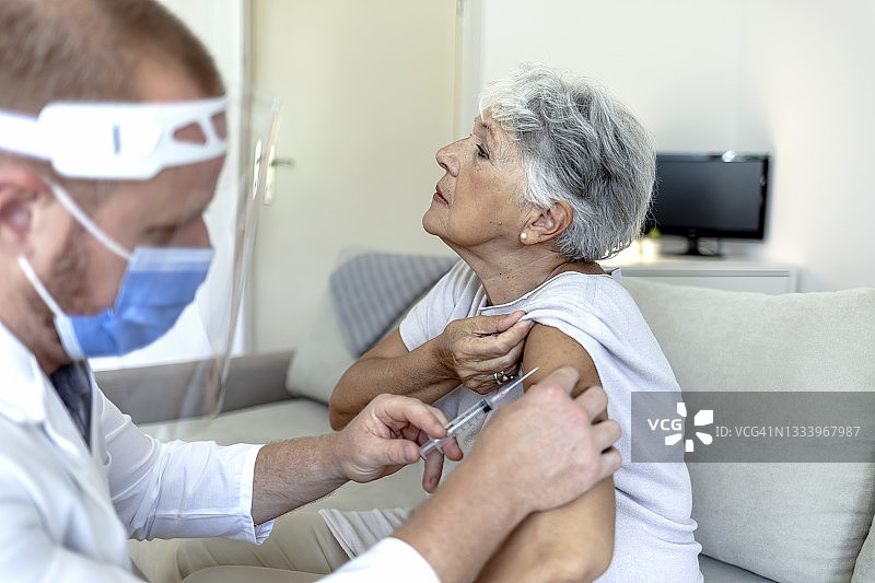 在冠状病毒爆发期间，戴着防护口罩的护士在家里给一位老妇人注射。图片素材