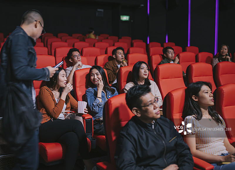 一名亚洲华人男子在电影放映时间进入电影院，试图穿过其他观众到达他的座位图片素材
