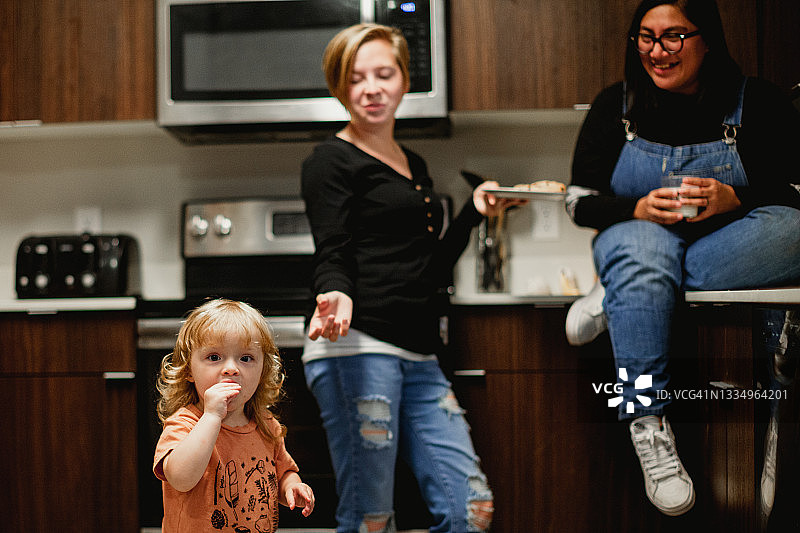 两个成年妇女和蹒跚学步的女孩在家庭厨房一起享受牛奶和饼干图片素材