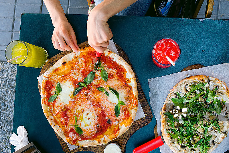 在户外的咖啡馆或快餐店里，一名女子用马苏里拉奶酪、番茄、罗勒切了一块素食披萨。美味可口的意大利菜。切成薄片的披萨玛格丽塔在木板或架子上。美味不健康的午餐，零食。图片素材