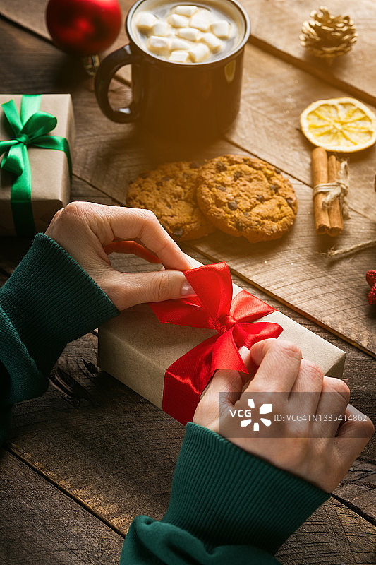 一个穿绿色毛衣的女孩或妇女拿着并系上圣诞礼物。全家准备过节的概念，平安夜快乐。木桌的背景上放着热咖啡和棉花糖，还有巧克力饼干。图片素材
