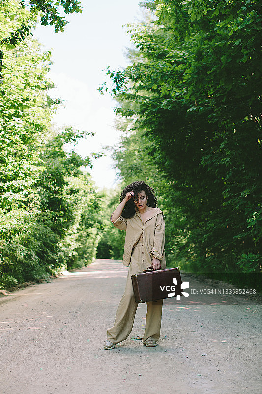 一个年轻的白色黑发女人，留着非洲式的发型，穿着米色的休闲服，拿着复古的手提箱站在路中间，对着镜头摆姿势，地平线被两旁的树木包围，变成了一个绿色的拱门。图片素材