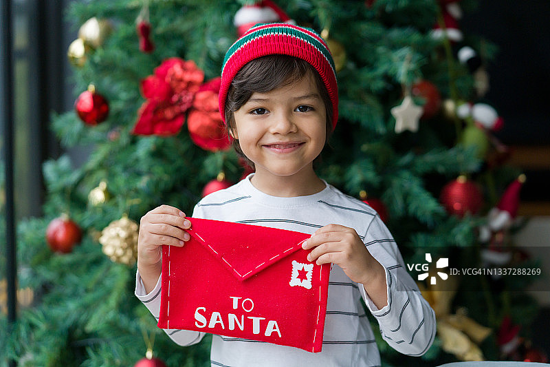 快乐的男孩手里拿着给圣诞老人的信，站在圣诞树旁图片素材