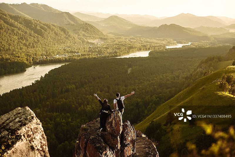 年轻快乐的徒步旅行者背着背包，从山顶举起双手，看和享受山谷和山间河流的景色。旅游度假概念图片素材