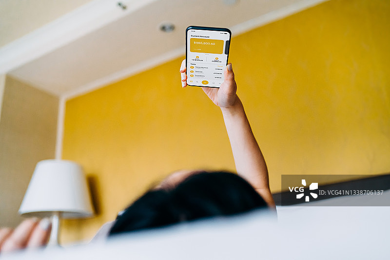年轻的亚洲女性在舒适的家里躺在床上放松，用智能手机上的移动应用管理网上银行。跟踪和计划支出。转账，支付账单，查看账户余额。在日常生活中使用科技的智能银行图片素材