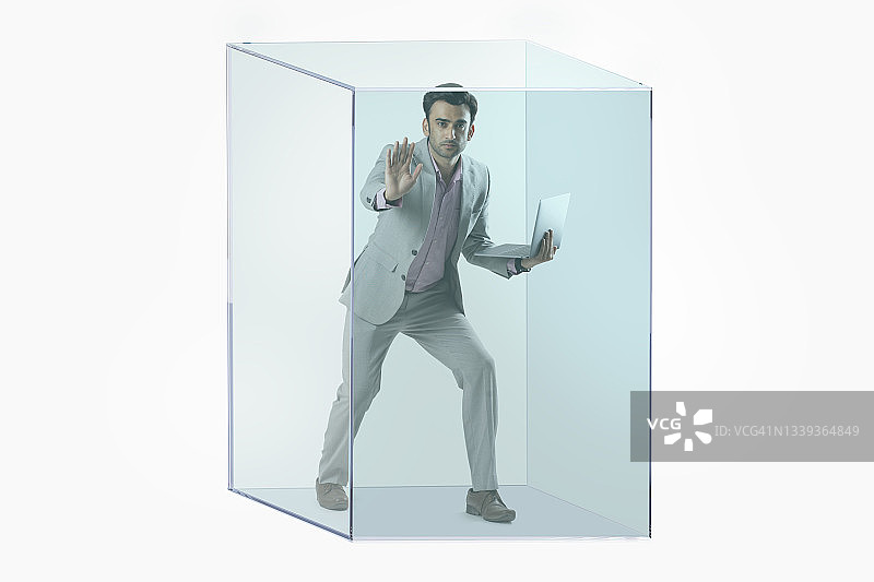 一名穿着正装的男子的肖像，他手里拿着一台笔记本电脑，被困在一个透明的立方体里。图片素材