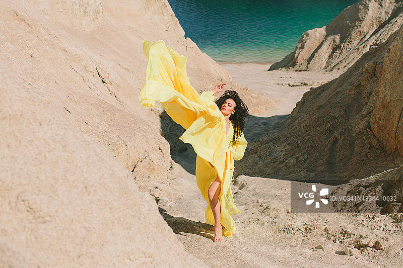一名年轻女子站在采石场的岸边，靠近沙石和湖泊，身穿黄色夏装，对着镜头摆姿势图片素材