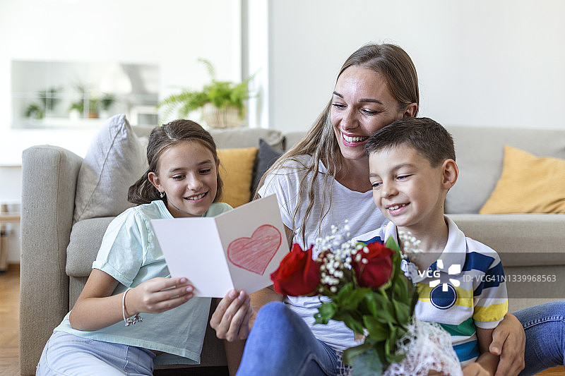 年轻的母亲抱着她的儿子笑着，在厨房庆祝节日的时候，活泼的女孩拿着卡片和玫瑰祝贺妈妈图片素材