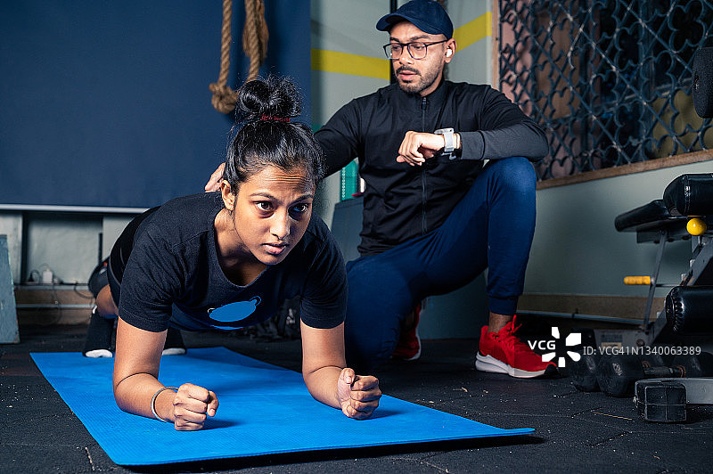 男教练帮助年轻女子在健身房做平板支撑练习。图片素材