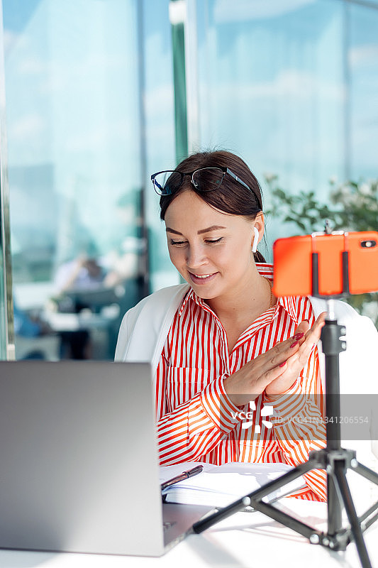 一个夏天，年轻的黑发女商人戴着眼镜坐在咖啡馆的笔记本电脑屏幕前，通过智能手机进行视频电话会议。业务、电子学习、自由的概念图片素材