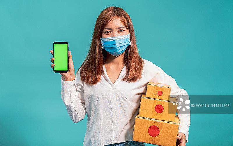 亚洲年轻女性在感染新冠病毒或COVID-19的情况下，身穿衬衫和戴着口罩的防护显示智能手机前绿色屏幕图片素材