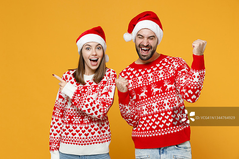 兴奋的年轻圣诞老人夫妇朋友，男人，女人在红色毛衣圣诞帽的食指在一边做胜者的手势孤立在黄色背景。新年庆祝快乐节日概念。图片素材