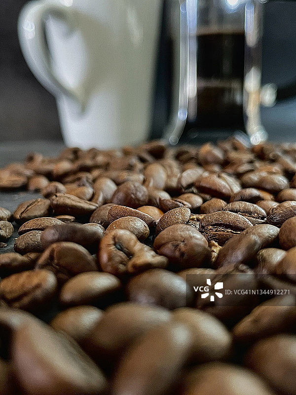 在白色咖啡杯和装满热黑咖啡的玻璃咖啡柜前的表面上烘焙咖啡豆的特写图像，逐渐缩小的视角，聚焦前景图片素材
