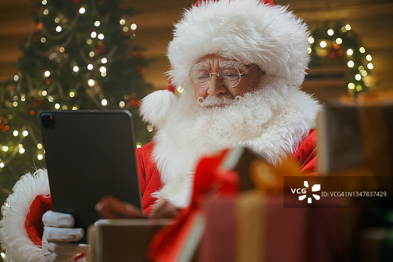圣诞老人坐在装饰着圣诞彩灯和圣诞树的办公室里，正全神贯注地看着手中的数码平板电脑，前景是礼物盒图片素材