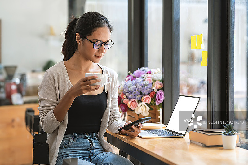 在café网站，亚洲女性戴着眼镜，一边喝咖啡一边使用智能手机的正面照片。图片素材