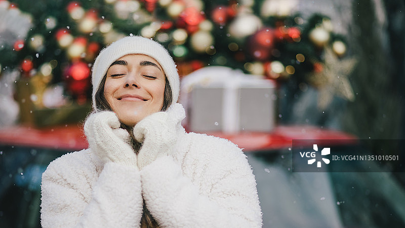 快乐美丽的年轻女子穿着针织毛衣和羊毛帽站在红色的汽车旁边，顶部有圣诞树在下雪。图片素材