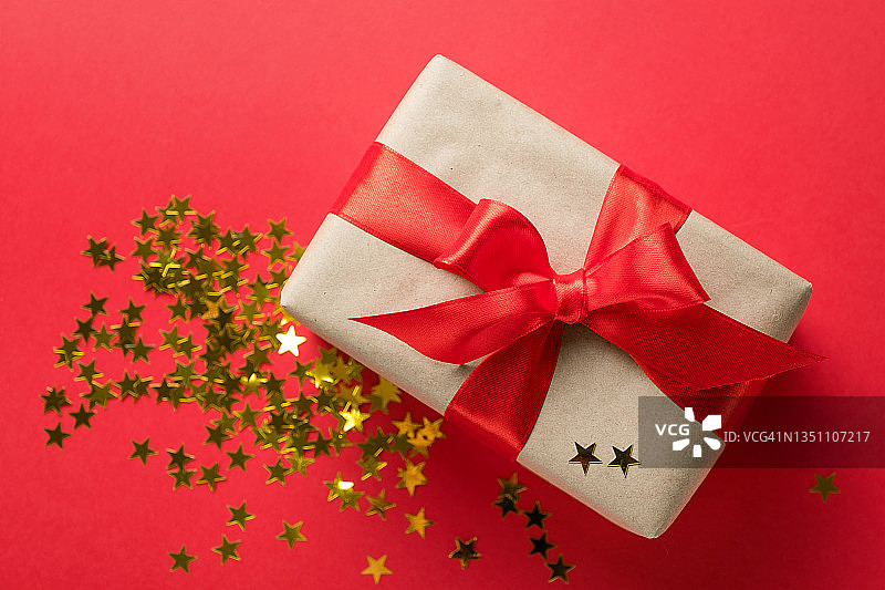 一个惊喜的包裹，用牛皮纸包裹，用鲜亮的丝带系在红色的背景上。附近散落着五彩纸屑，形成闪烁的金色星星。在节日前夕互赠礼物。快乐圣诞节的概念。图片素材