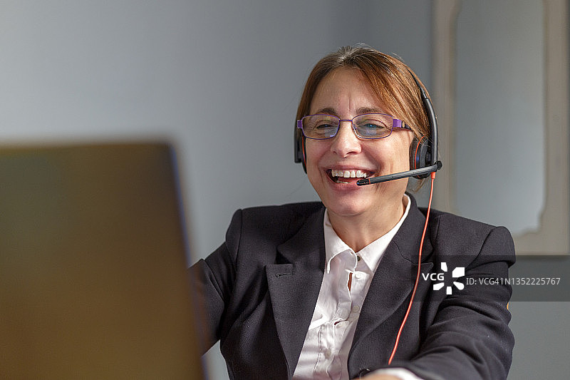 在呼叫中心，一位戴着眼镜、耳机和麦克风微笑的中年妇女看着笔记本电脑屏幕。马德里,西班牙图片素材