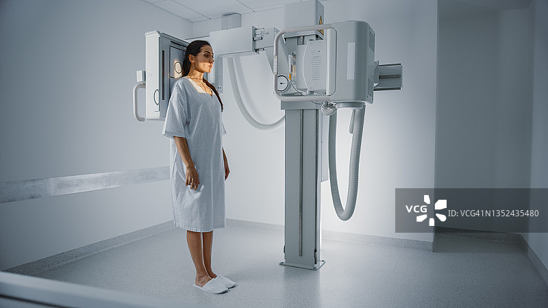 医院放射科:美丽的多民族妇女穿着医用长袍站在x光机上。成年女性在现代诊所接受健康检查，正在扫描胸、心、肺。图片素材