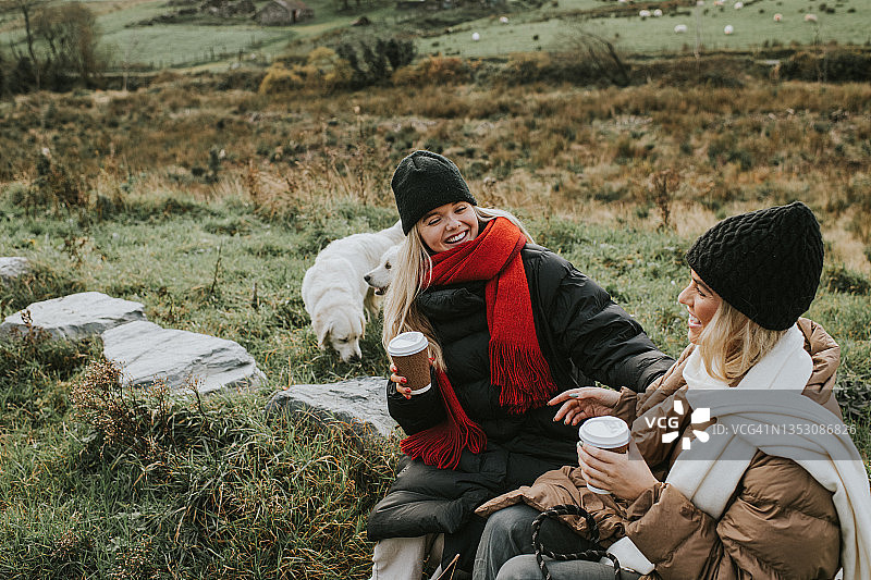 两个活泼、漂亮的年轻女子坐在大石头上，手里拿着外卖杯，裹着暖和的外套、围巾和帽子，聊着天。两只金毛猎犬站在背景中。图片素材