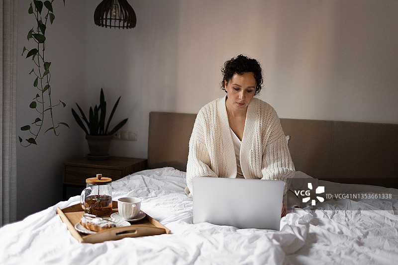 这位女商人在家工作，坐在一张白色的床上，一边用笔记本电脑聊天，一边吃早餐图片素材