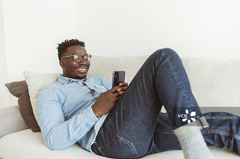 懒懒的非洲人穿着休闲服在现代温暖的客厅里拿着智能手机和朋友聊天。图片素材