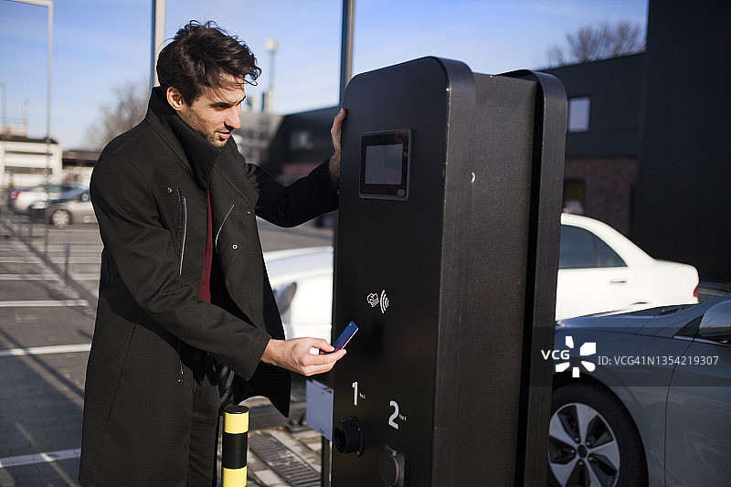 一个年轻人在停车场，用信用卡为他的电动汽车充电图片素材