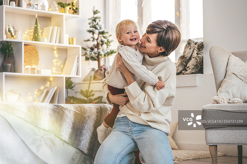 在一间装饰成圣诞节的房间里，年轻的母亲抱着她微笑的蹒跚学步的婴儿图片素材