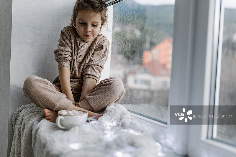 一个穿着白色针织睡衣的小女孩坐在窗边，用杯子喝水图片素材
