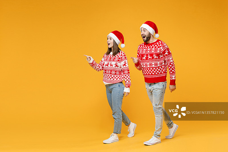 全身震惊的年轻圣诞老人夫妇的朋友，男人女人穿毛衣，圣诞帽点食指在一旁孤立的黄色背景工作室肖像。欢度新年的节日理念。图片素材
