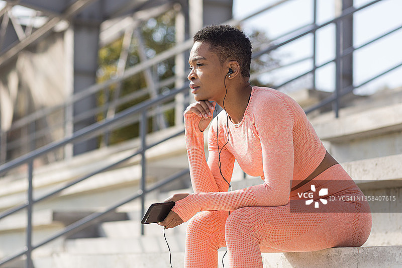 严肃的年轻女运动员在锻炼后穿着粉红色的西装坐着休息，用手机听音乐和播客图片素材