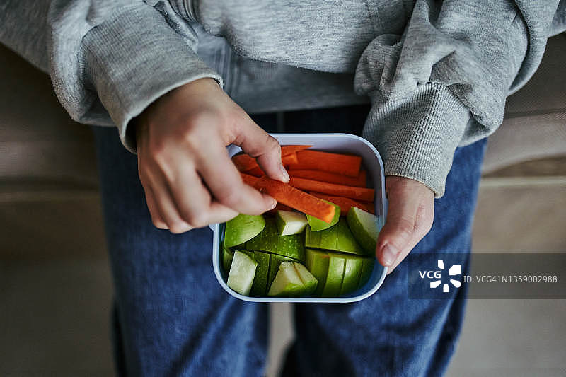一个孩子手里拿着一个午餐盒，里面有健康的食物，坚果，胡萝卜和一个苹果。图片素材